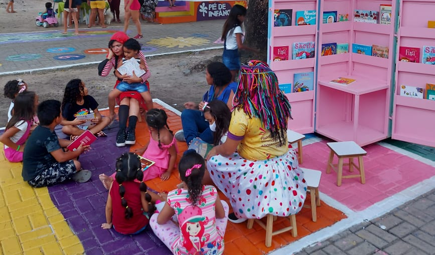 na foto, crianças estão sentadas em volta do quiosque com livros